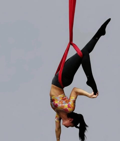 Aerial performer doing an inverted handstand while suspended from a red fabric sling against a gray background