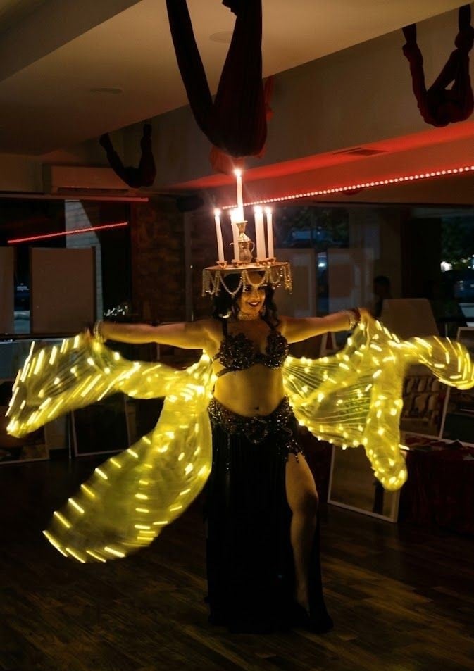 Performer in glowing yellow LED wings and black sequined costume posing with arms outstretched in an illuminated venue with red lighting