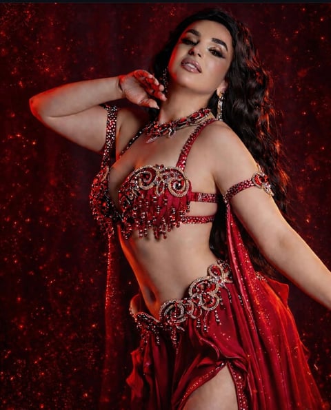 Woman wearing ornate red beaded belly dance costume with matching jewelry and dramatic makeup against a glittering red background