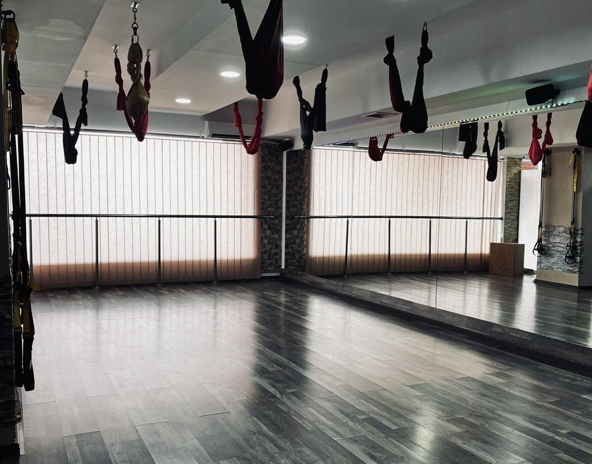 Aerial yoga studio with suspended silks and hammocks hanging from ceiling, polished wood floor, large windows with blinds, and mirror wall