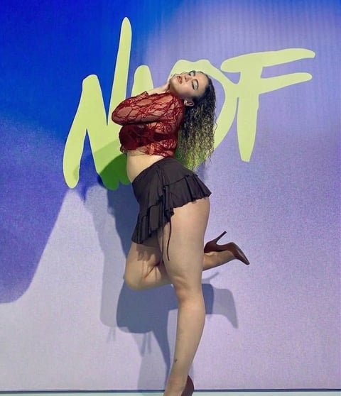 Person with long red wavy hair wearing black crop top and skirt, posing against colorful gradient background with yellow graffiti-style text