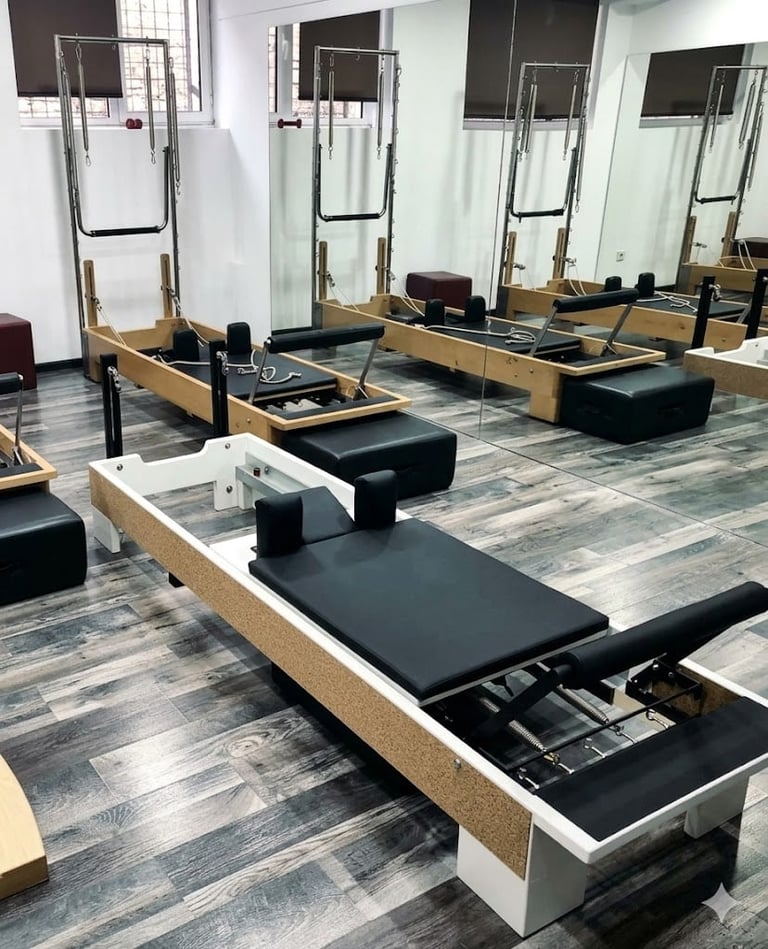 A spacious Pilates studio with rows of wooden reformer machines with black padding, mirrors, and bright windows